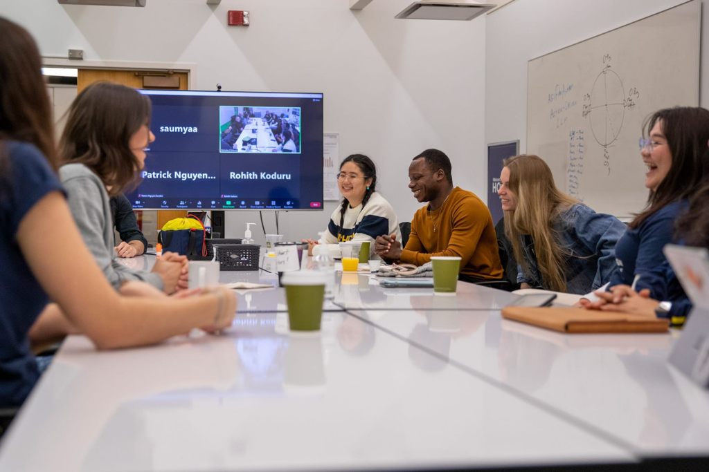Design Science students smiling and collaborating around a table during a hybrid class meeting.